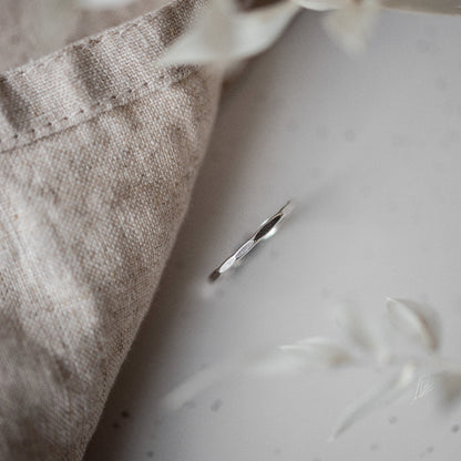 A simple, elegant silver stacking ring with a hammered finish, displayed on a neutral background with dried plants beside it.