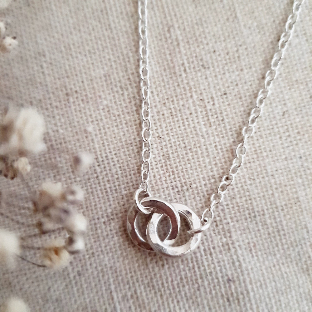 A silver necklace with two linked hammered silver circles as the central pendant, displayed on a textured background.