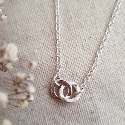 A silver necklace with two linked hammered silver circles as the central pendant, displayed on a textured background.