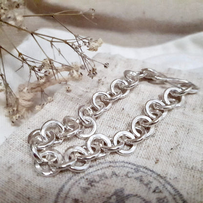 A chunky hammered link silver bracelet laid out next to some dried flowers on a textured fabric surface.