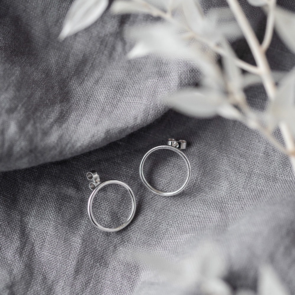 A pair of large silver circle stud earrings placed on a light grey fabric with blurred white leaves.