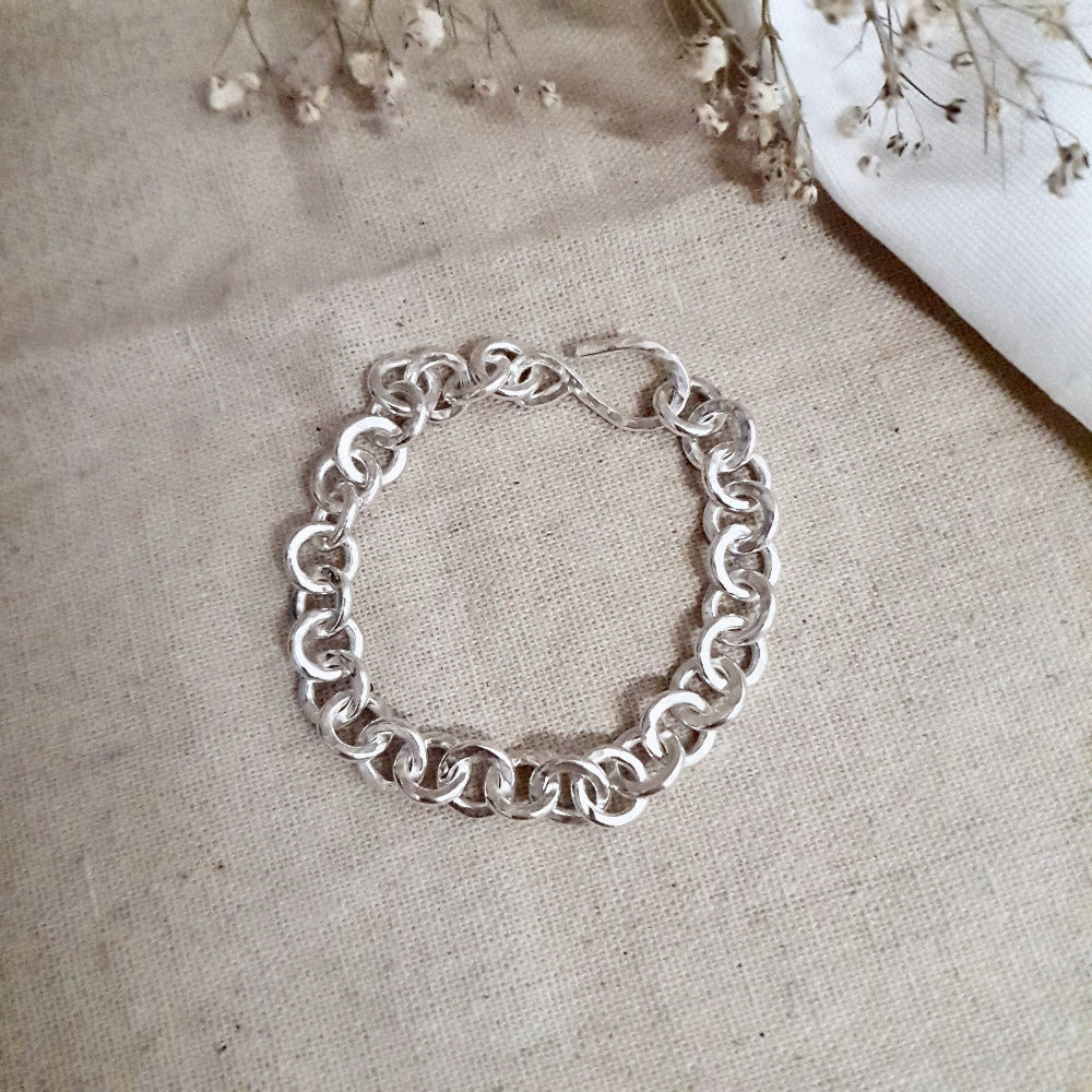 A chunky hammered link silver bracelet laid out next to some dried flowers on a textured fabric surface.