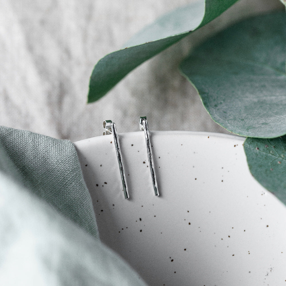 A pair of hammered silver bar earrings hanging from the edge of a ceramic dish with leaves in the background