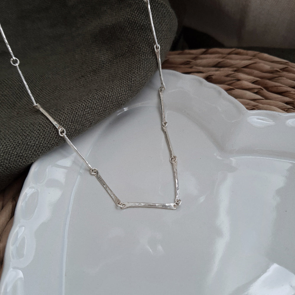 A silver bar chain necklace with a hammered texture, displayed on a green textured background and white plate
