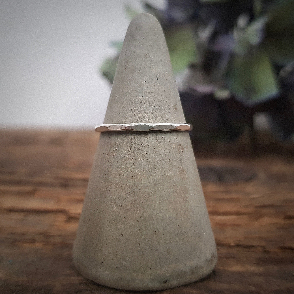 A simple, elegant silver stacking ring with a hammered finish, displayed on a grey ring cone with dried plants behind it.