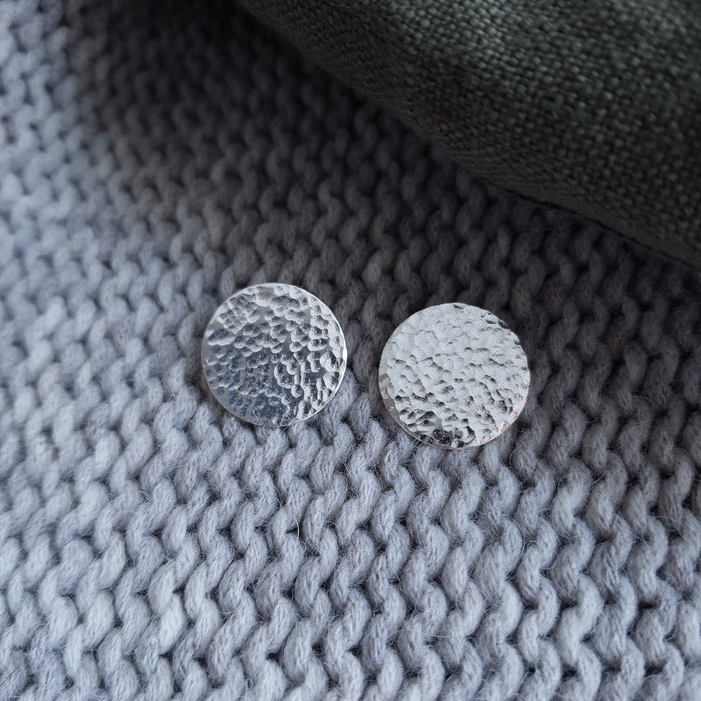 A pair of silver hammered disc earrings resting on a  grey woven surface.