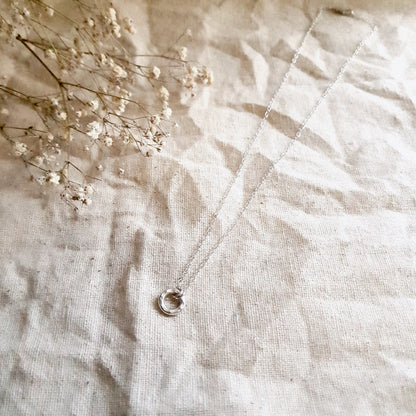A sterling silver necklace with a small hammered circular pendant, displayed on a white fabric background.