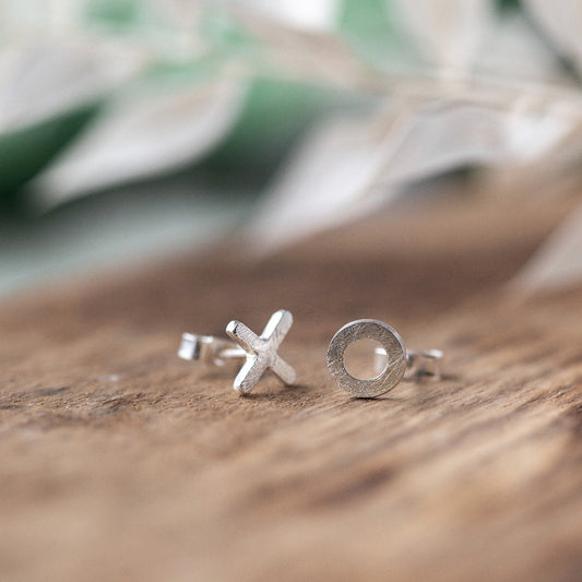 A pair of silver stud earrings with a textured finish, one is a circle the other a X placed on a wooden surface with a blurred green leafy background.