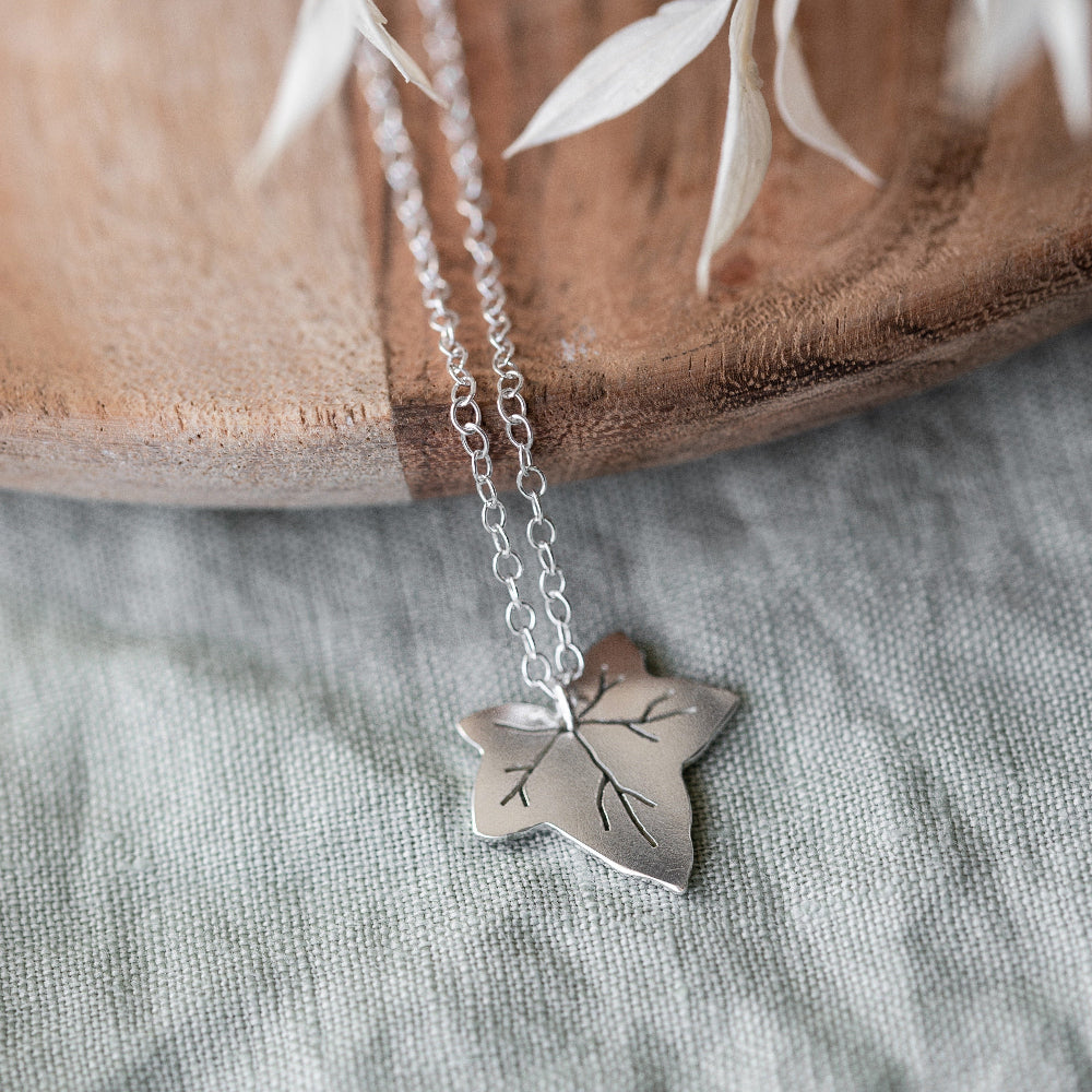 A silver necklace featuring a Ivy leaf-shaped pendant, displayed against a neutral background and a piece of wood