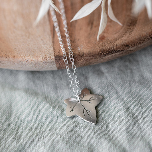 A silver necklace featuring a Ivy leaf-shaped pendant, displayed against a neutral background and a piece of wood