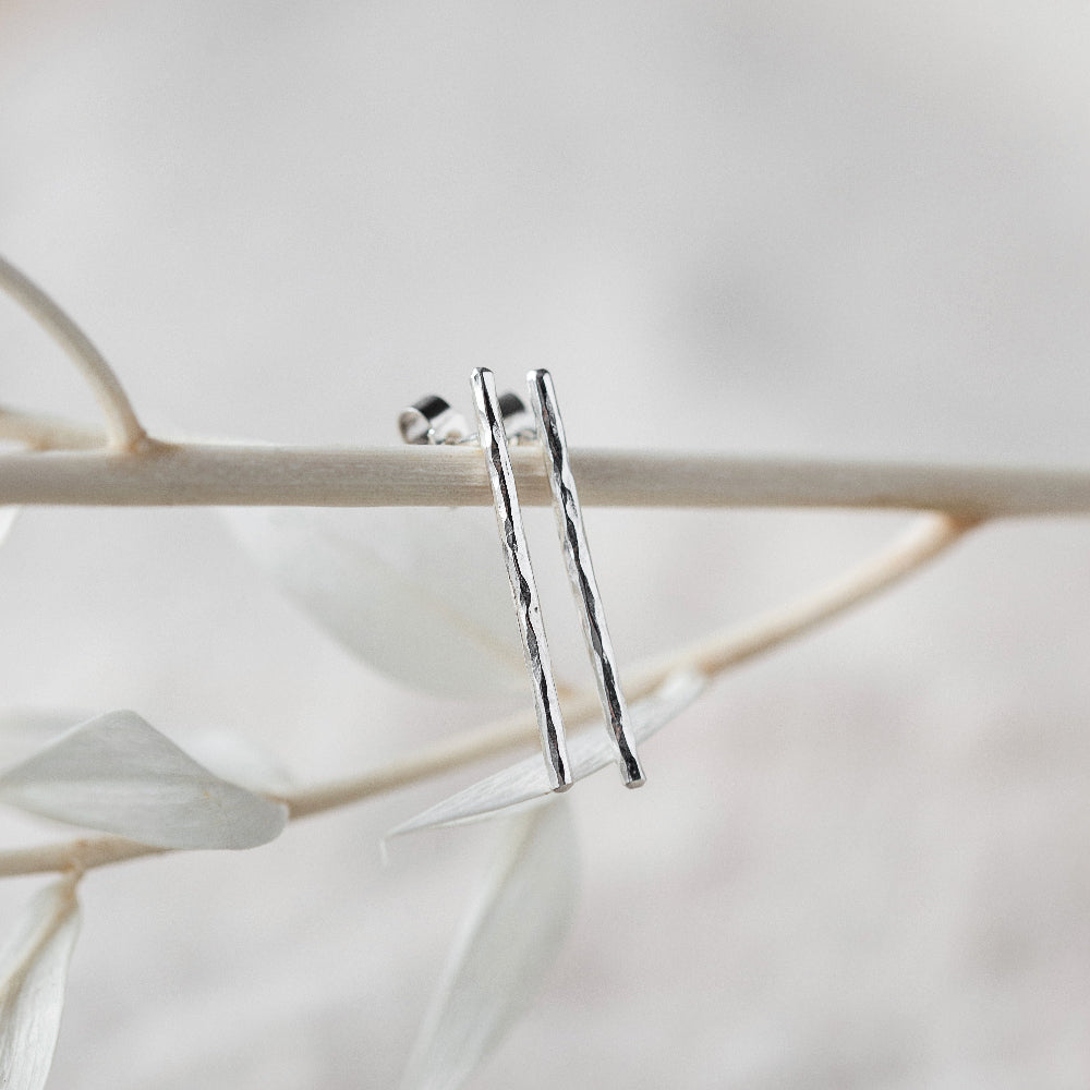 A pair of hammered silver bar earrings hanging from a branch with a blurred background.