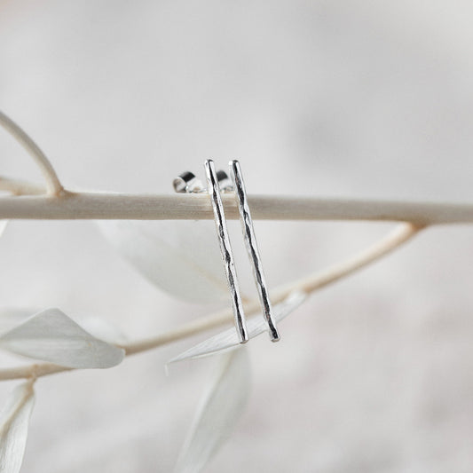 A pair of hammered silver bar earrings hanging from a branch with a blurred background.