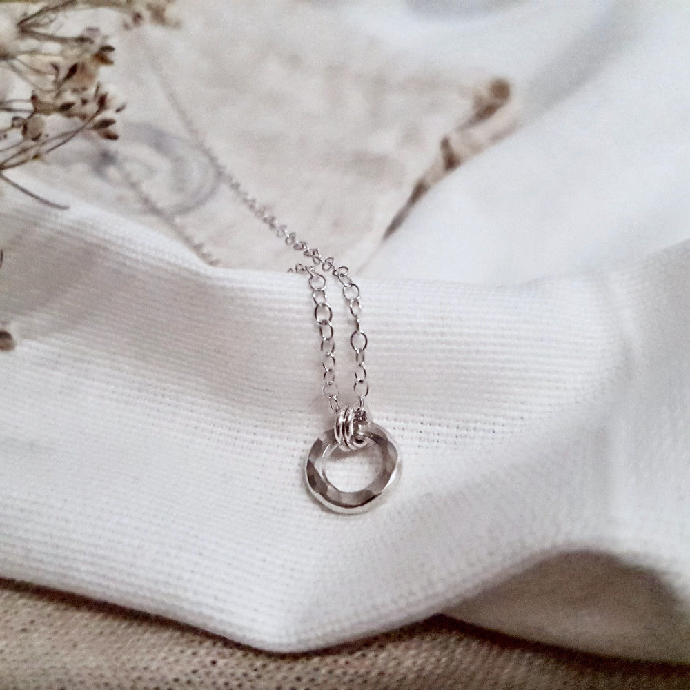 A sterling silver necklace with a small hammered circular pendant, displayed on a white fabric background.