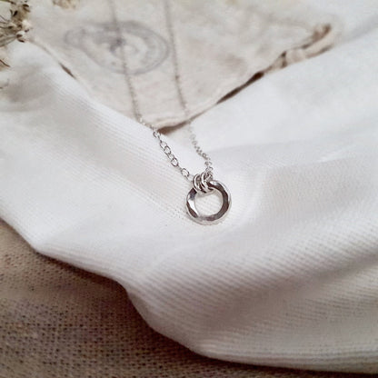 A sterling silver necklace with a small hammered circular pendant, displayed on a white fabric background.