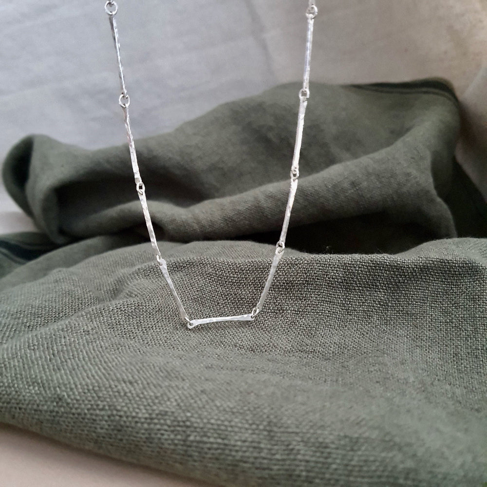 A silver bar chain necklace with a hammered texture, displayed on a green textured background.
